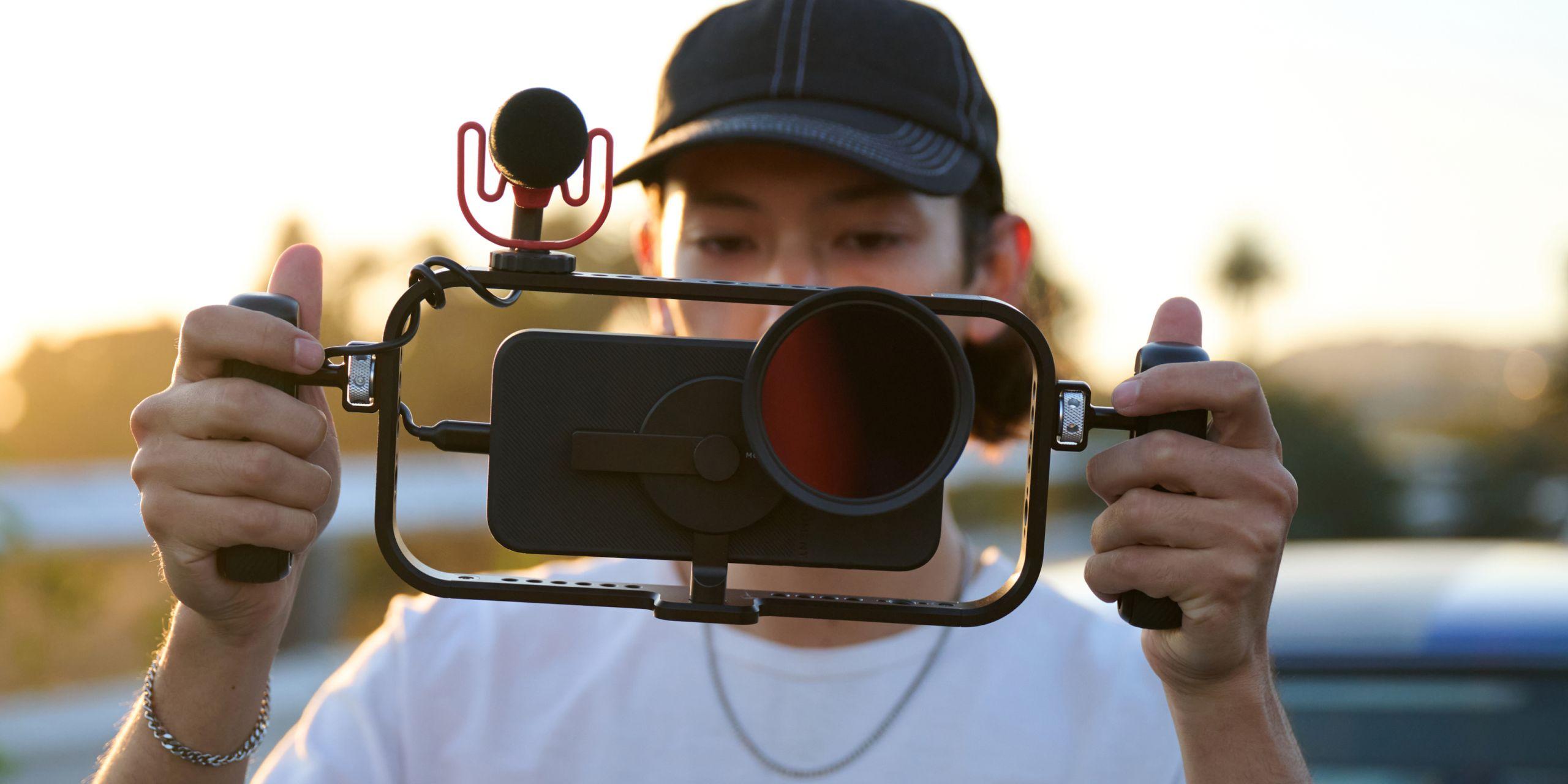 Hand holding iPhone 16 with a mobile filmmaker cage with an on-camera shotgun microphone for footage.